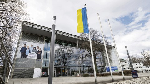 Das Foto zeigt das Hörsaalgebäude der TU Dresden. Davor stehen drei Fahnenmasten, an dem linken ist die Flagge der Ukraine gehisst.