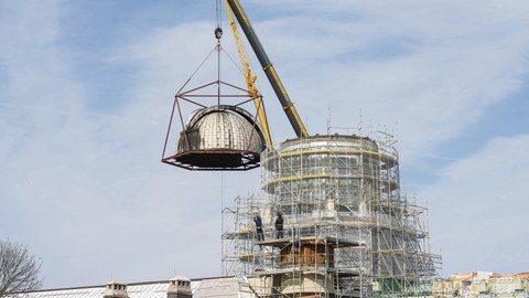 Zu sehen ist der eingerüstete Turm des Beyer-Baus. Auf dem Gerüst stehen zwei Menschen. An einem Kran hängt die Kuppel.