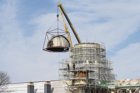 Zu sehen ist der eingerüstete Turm des Beyer-Baus. Auf dem Gerüst stehen zwei Menschen. An einem Kran hängt die Kuppel.