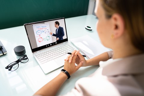 Foto einer Person, die an ihrem Schreibtisch sitzt und auf ihrem Laptop eine Online-Veranstaltung verfolgt.