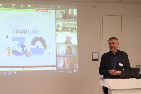 Prorektor Bildung am Pult stehend vor Leinwand mit Präsentation und Bildern von Online-Teilnehmenden 