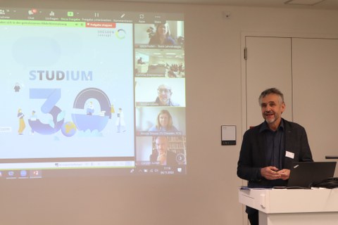 Prorektor Bildung am Pult stehend vor Leinwand mit Präsentation und Bildern von Online-Teilnehmenden