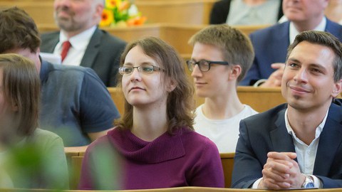 junge und ältere Menschen in guter Kleidung sitzen vergnügt in den Reihen eines Hörsaals.