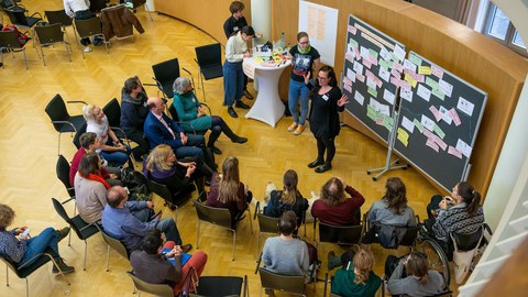 Luftaufnahme von circa 20 Menschen, die im Dülfersaalv im Halbrund sitzen und einer Frau, vor einer Tafel stehend, zuhören.