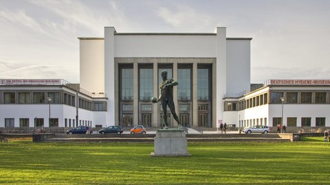 Quadratisches Gebäude mit Vorplatz und der Statue eines Sportlers