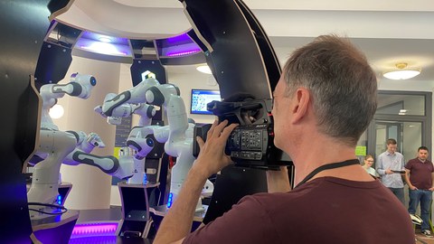 ein Kameramann schaut durch eine Kamera und filmt dabei drei interagierende Roboterarme
