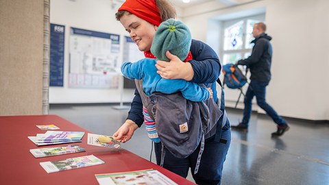 kleuinwüchsige Frau mit Baby steht an einem Tisch mit Infomaterial
