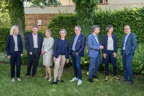 Group photo of the University Executive Board. All eight members are standing in front of a green hedge on a meadow, talking and laughing with each other.