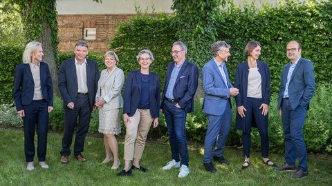 Group photo of the University Executive Board. All eight members are standing in front of a green hedge on a meadow, talking and laughing with each other.