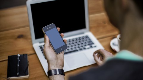 Mann sitzt mit Smartphone vor Laptop