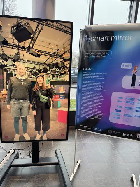 Two people stand in front of a large screen showing their full-size reflection with studio lighting and equipment in the background, next to an informational poster about a smart mirror with text and graphics.