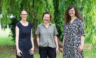 Stephanie Feilitzsch, Jutta Eckhardt, and Marie Kukuczka are standing next to each other outdoors in front of the hanging branches of a tree. In the background, a green meadow is visible. Stephanie Feilitzsch is on the left. She is wearing a dark, short-sleeved dress and glasses. Jutta Eckhardt is in the middle. She is wearing a short-sleeved, light shirt and dark trousers. Marie Kukuczka is on the right. She is wearing a patterned, long dress and glasses. All three are smiling and looking toward the camera.