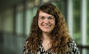 Porträtfoto Marie Kukuczka. Eine Frau mit langen, lockigen braunen Haaren und Brille lächelt in die Kamera. Sie trägt eine gemusterte Bluse in Schwarz und Beige. Im Hintergrund sind große Fenster mit unscharfem Blick ins Grüne zu sehen.