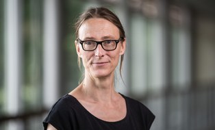 Porträtfoto Dr. Stephanie Feilitzsch. Eine Person mit langen, zurückgebundenen Haaren und Brille steht in einem Flur mit unscharfem Hintergrund. Sie trägt ein dunkles Oberteil und blickt ruhig in die Kamera.