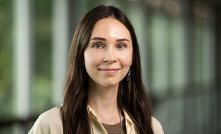 Porträtfoto Maryna Havriushyna. Eine Person mit langen, dunklen Haaren steht in einem Flur mit unscharfem Hintergrund. Sie trägt eine helle Bluse über einem braunen Oberteil, blickt direkt in die Kamera und lächelt leicht.