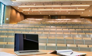 Das Foto zeigt einen leeren Hörsaal. Im Vordergrund erkennt man ein Heft und einen Laptop auf dem Rednerpult.