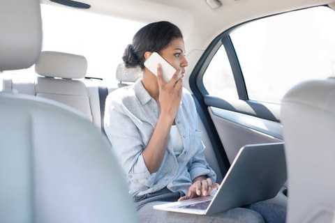 Foto einer Person im Auto. Sie sitzt auf dem Rücksitz und hält ein Mobiltelefon in der Hand. Auf ihrem Schoß liegt ein aufgeklappter Laptop.