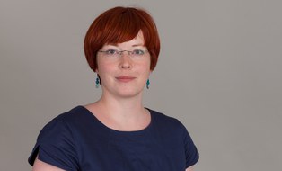 Porträtaufnahme von Carola Queitsch. Wir sehen eine mitteljunge, lächelnde Frau mit Brille und roten Haaren in grünem Oberteil und grüner Jacke vor einem unscharfen, lichten grünen Hintergrund.