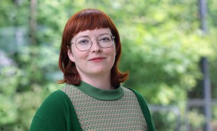 Porträtaufnahme von Carola Queitsch. Wir sehen eine mitteljunge, lächelnde Frau mit Brille und roten Haaren in grünem Oberteil und grüner Jacke vor einem unscharfen, lichten grünen Hintergrund.