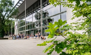 The photo shows the exterior view of the TU Dresden lecture hall centre. There are many people in front of the lecture hall centre. In the foreground you can see the leaves of a tree.