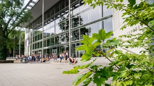 Das Foto zeigt die Außenansicht des Hörsaalzentrums der TU Dresden. Vor dem Hörsaalzentrum befinden sich viele Personen. Im Vordergrund erkennt man die Blätter eines Baumes.