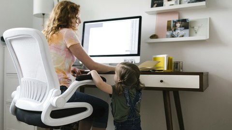 Foto zeigt eine Mutter im Homeoffice am Computer, die nebenbei mit ihrer kleinen Tochter spricht.