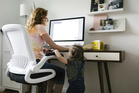 Foto zeigt eine Mutter im Homeoffice am Computer, die nebenbei mit ihrer kleinen Tochter spricht.