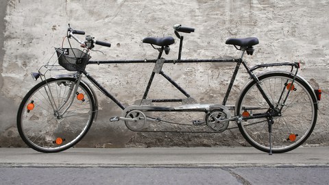  (ein Fahrrad für zwei Personen). Das Tandem steht vor einer Hauswand.