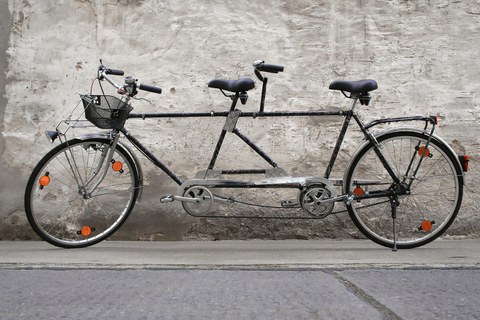  (ein Fahrrad für zwei Personen). Das Tandem steht vor einer Hauswand.