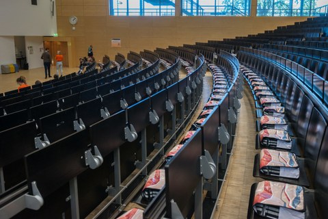 Das Foto zeigt die leeren Bankreihen eines Hörsaals. Dabei liegt jedoch auf jedem der Stühle eine gefüllte Umhängetasche.