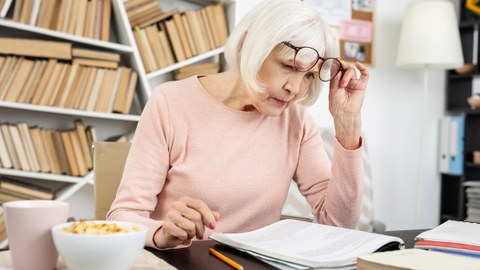 Eine ältere Dame sitzt mit konzentriertem Blick an einem Arbeitstisch und liest in einem Dokument.