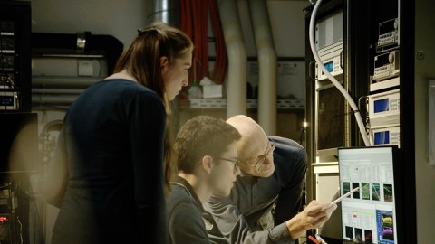 Three scientists are looking at a screen. One points at the screen with a pencil.