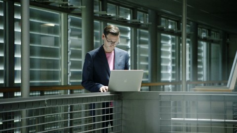Prof. Koch ist im rechten Teil des Fotos. Er steht an einem Geländer, auf dem ein aufgeklappter Laptop steht, in den er hineinblickt. Im Hintergrund geschlossene Außenjalousien.