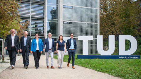 The six Speakers of the Clusters of Excellence - Professors Fitzek, Vojta, Vaynzof, Mechtcherine, von Kriegstein and Campàs - walk side by side on the lawn behind the Auditorium Center. The TUD logo is visible in the background.