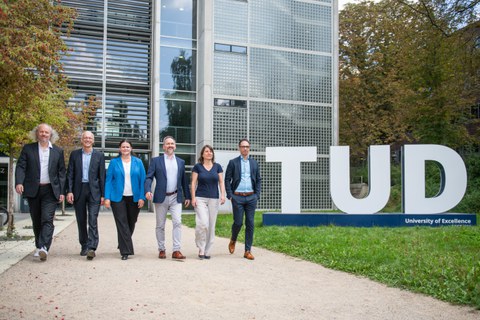 The six Speakers of the Clusters of Excellence - Professors Fitzek, Vojta, Vaynzof, Mechtcherine, von Kriegstein and Campàs - walk side by side on the lawn behind the Auditorium Center. The TUD logo is visible in the background.