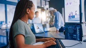 Eine fokussierte, junge Frau mit Zöpfen und einem grünen T-Shirt tippt auf einer Tastatur. Sie blickt auf zwei Monitore, auf denen Computercode und ein 3D-Modell einer Roboterhand zu sehen sind. Im Hintergrund interagieren zwei weitere Personen.