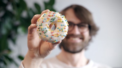 Mann im verschwommenen Hintergrund hält einen Donut in die Kamera