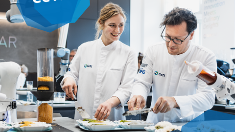 Foto von zwei Personen beim Zubereiten von Essen an einem Tresen. Darauf steht ein Roboter und unterstützt beim Zubereiten der Mahlzeiten.