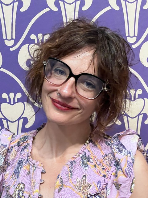 Prof. Orit Halpern in einer Bluse mit Blumenmuster vor einer expressionistischen Wandtapete