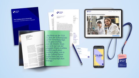 Mehrere Branding-Elemente der TU Dresden, darunter ein dunkelblaues Heft mit weißem Text, ein offenes Magazin mit grünem und weißem Text, ein Briefbogen, ein Stift, ein Smartphone mit der Zahl '29.000', ein Tablet mit einer Webseite und ein Schlüsselband.