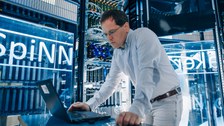 Foto: Prof. Christian Mayr lehnt sich auf einen Tisch mit Laptop, umgeben von Serverracks des Supercomputers SpiNNcloud.