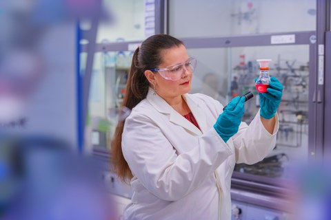 Foto: Professorin Yana Vaynzof trägt einen weißen Laborkittel und blaue Schutzhandschuhe. Sie hält ein Reagenzglas mit einer roten Flüssigkeit in der Hand. Im Hintergrund sind Laborgeräte und Regale sichtbar.