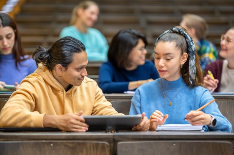 Foto: Studierende sitzen in einem Hörsaal, zwei sitzen sich zugewandt im Vordergrund mit Tablet und Notizblock, sind konzentriert beim Lernen.