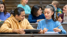 Foto: Studierende sitzen in einem Hörsaal, zwei sitzen sich zugewandt im Vordergrund mit Tablet und Notizblock, sind konzentriert beim Lernen.