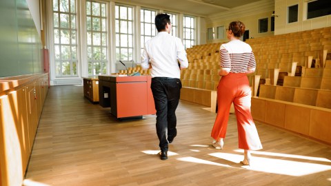 Foto: Zwei Dozenten gehen in einem hellen Hörsaal zum Sprechpult, sie sind von hinten zu sehen. Der Hörsaal hat große Fenster und eine Holzbestuhlung.