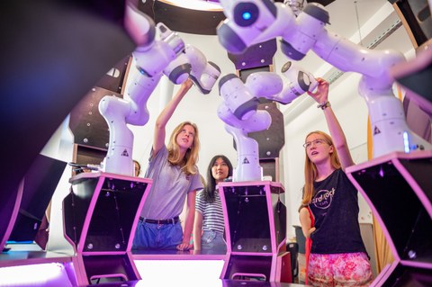 Schüler:innen stehen in einem Raum mit Roboterarmen, die rosa beleuchtet sind und zeigen darauf. 