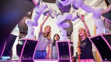 Schüler:innen stehen in einem Raum mit Roboterarmen, die rosa beleuchtet sind und zeigen darauf. 