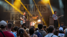 Foto: Drei Musiker spielen auf einer Bühne vor Publikum beim Campusfest mit gelben Bühnenlichtern
