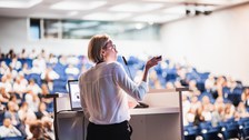 Foto: Eine Person spricht an einem Rednerpult vor einem Publikum in einem Konferenzraum