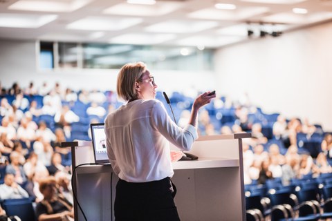 Foto: Eine Person spricht an einem Rednerpult vor einem Publikum in einem Konferenzraum